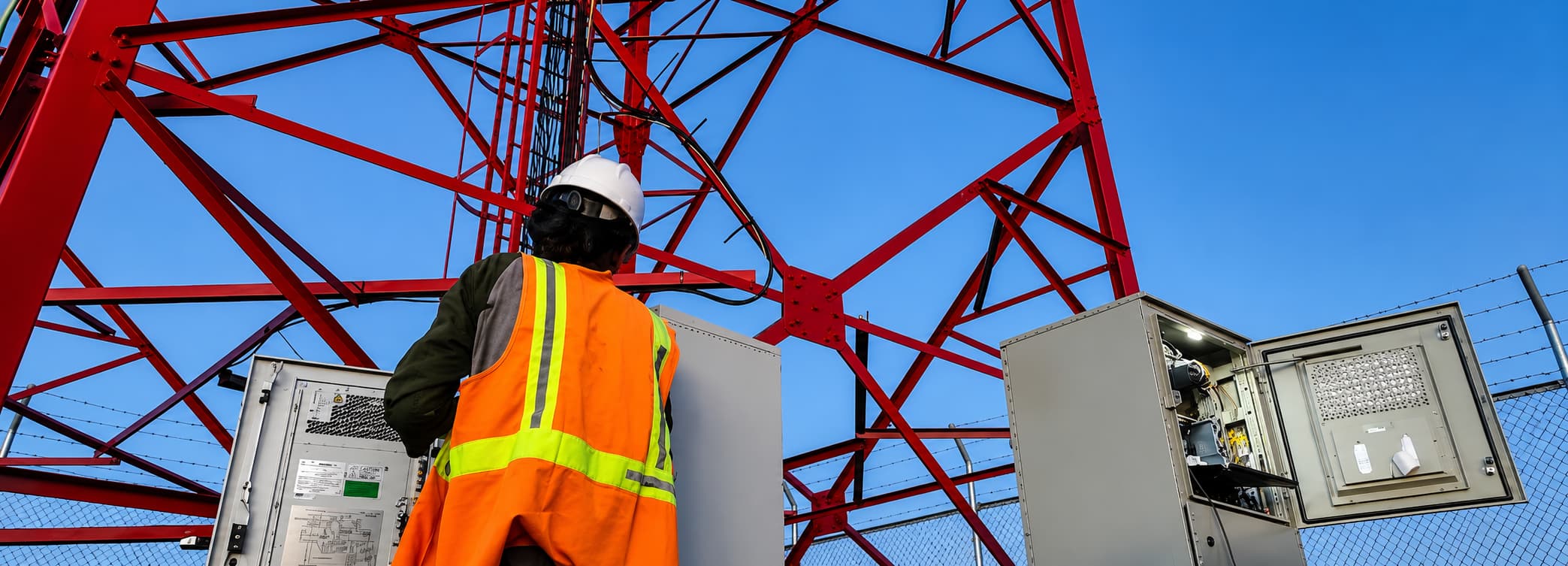 Field engineer wearing PPE working on an electrical control cabinet at the base of a telecoms mast, carrying out maintenance on network power infrastructure.